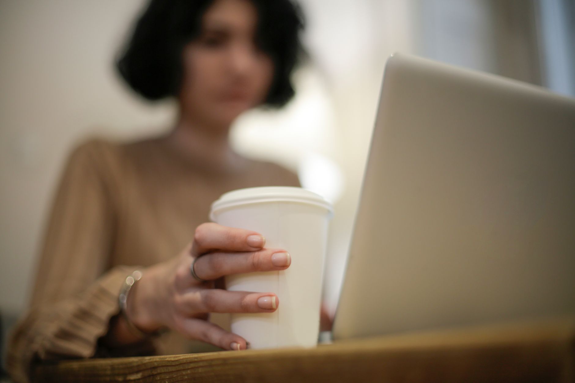 Una mujer desenfocada sostiene un vaso de café para llevar junto a un portátil abierto sobre una mesa de madera.